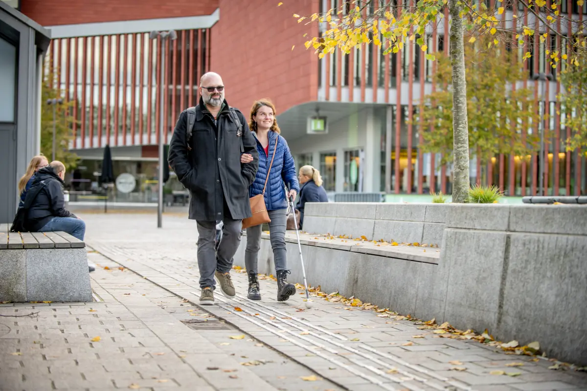 Blind woman with a white cane walks arm in arm with a male companion on tactile guidance strip on 't Zand Square