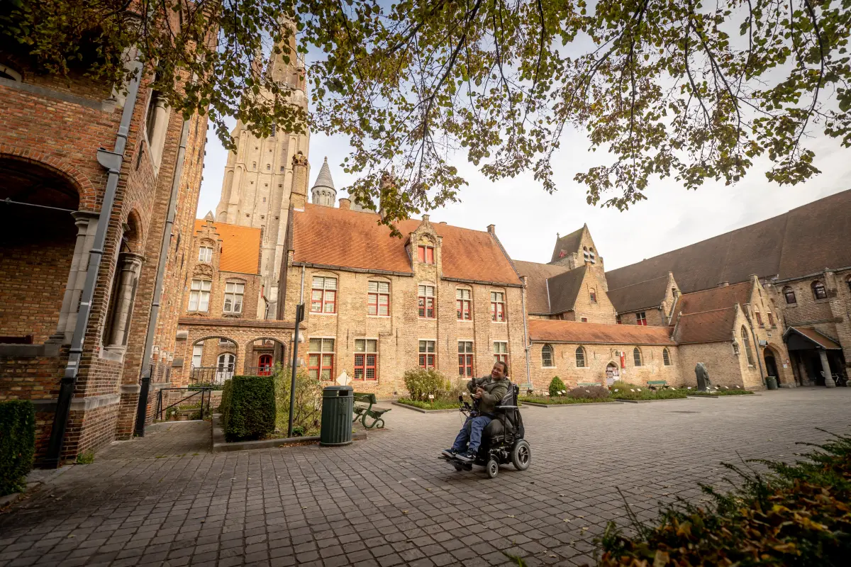 Eleonora Verbekehof at the Old St. John's site, where a man in an electric wheelchair is driving, with the St John’s Hospital and the tower of the Church of Our Lady behind him.