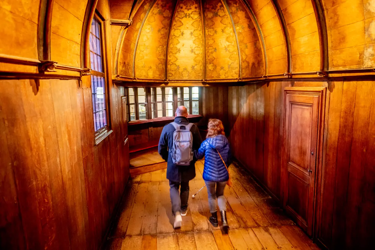 visually impaired visitor with an assistant in the authentic medieval prayer chapel in the Gruuthuse Museum, with a wooden floor, paneling, and a vaulted ceiling