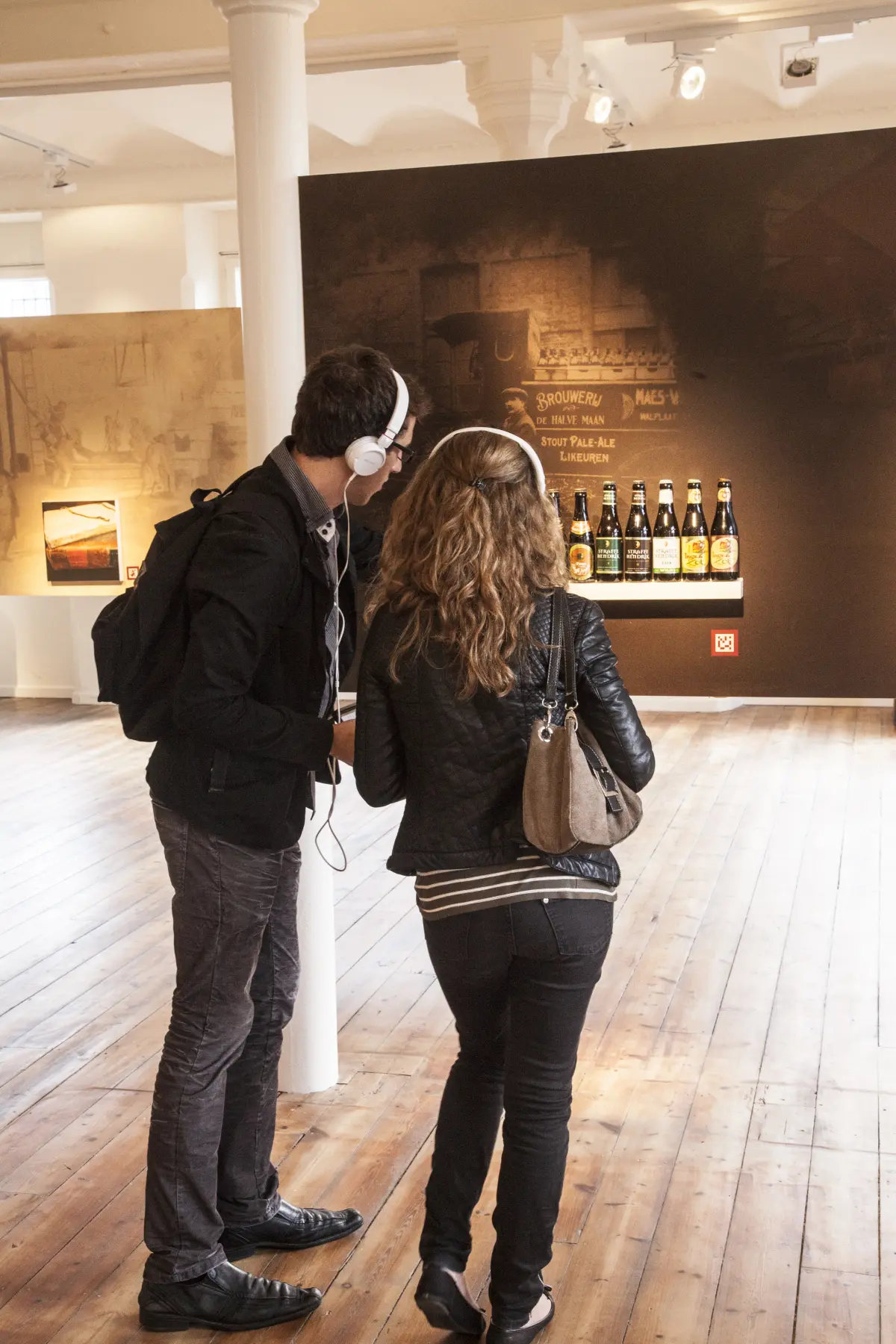 two visitors with an audio guide examining examples of beer bottles