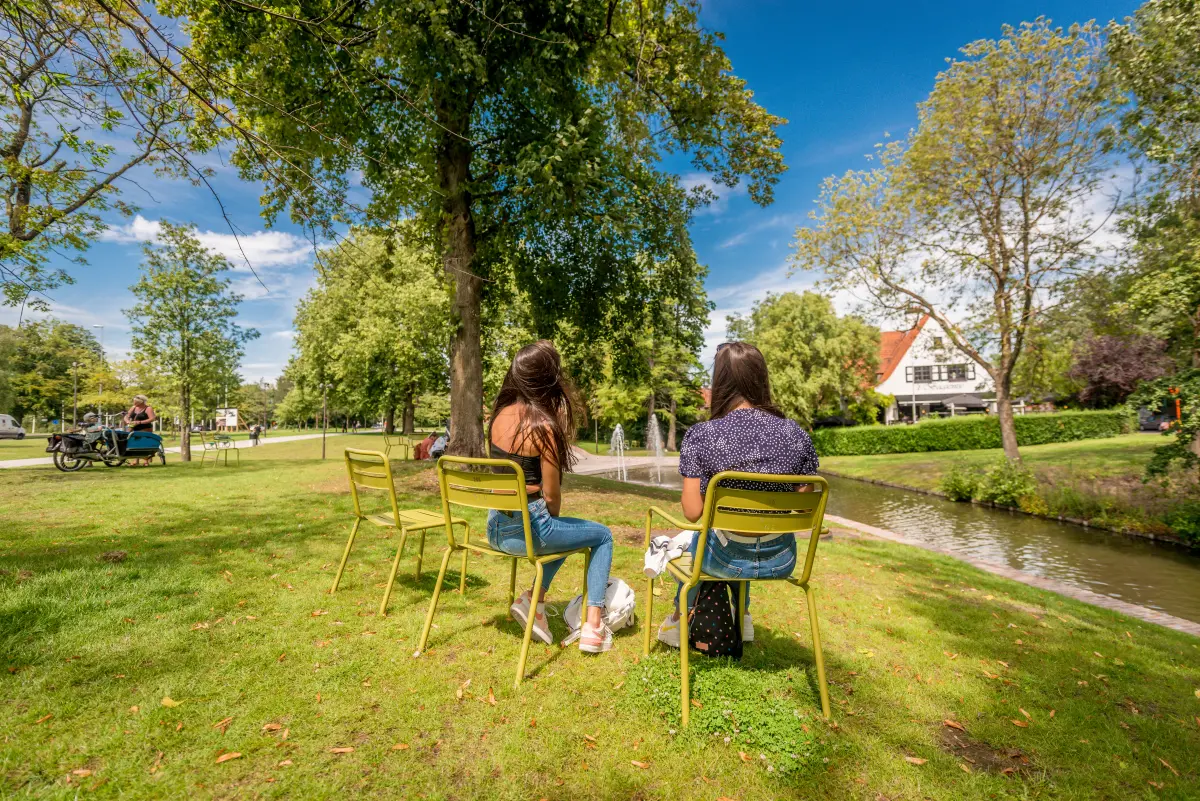 King Albert I park with two female visitors sitting on green chairs in the foreground and resting cyclists in the background.